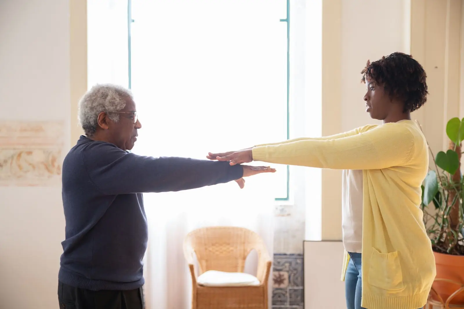 A care worker and an older man face each other with their arms stretched out, doing an exercise together in a bright room.