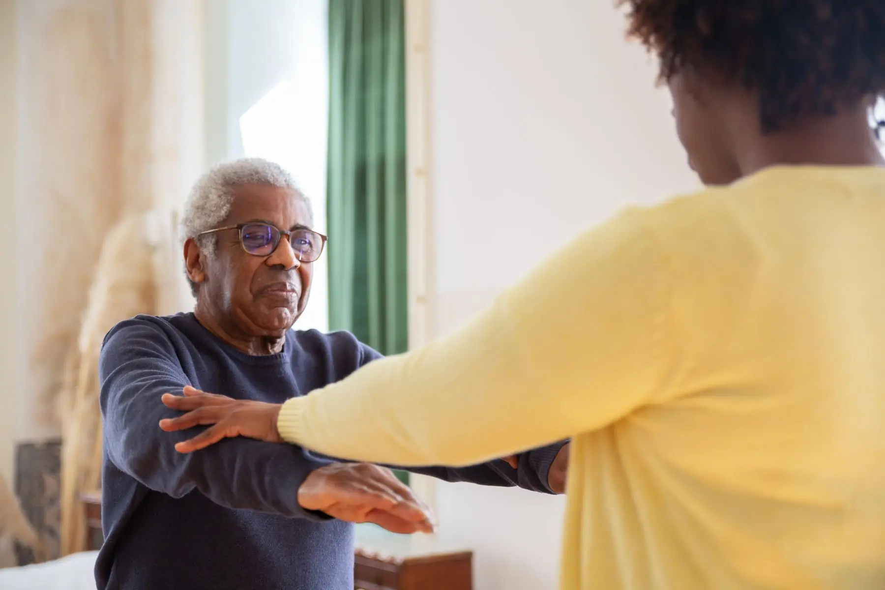 An older man with glasses holds both arms out in front of him while a care worker guides his exercise.
