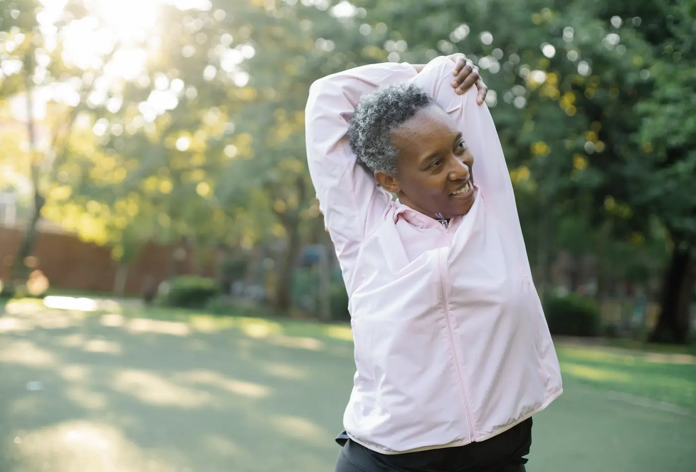 An older woman in a pink top stretches her arms overhead in a sunlit park.